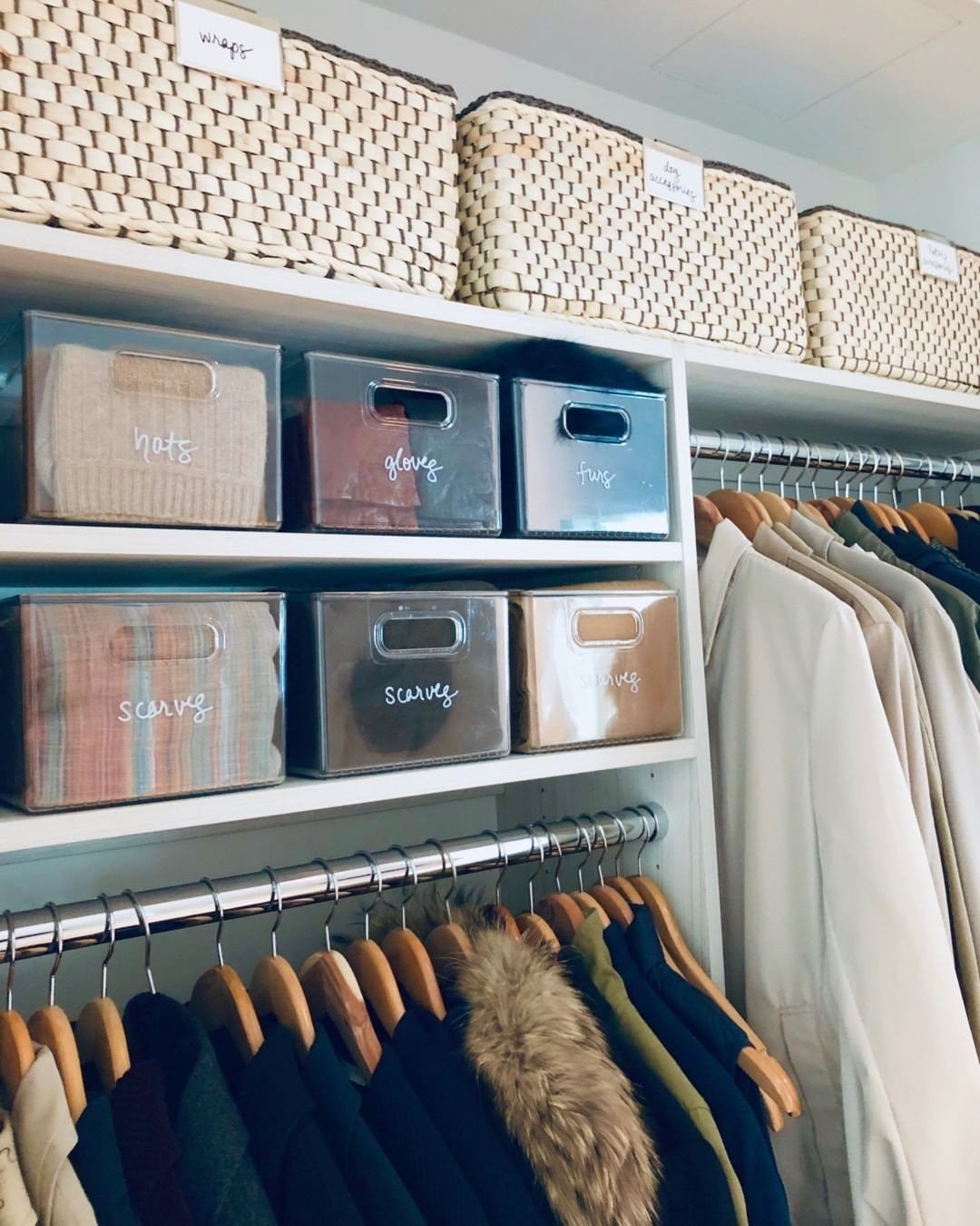 Clear plastic bins with labels on white closet shelves above hanging clothes.