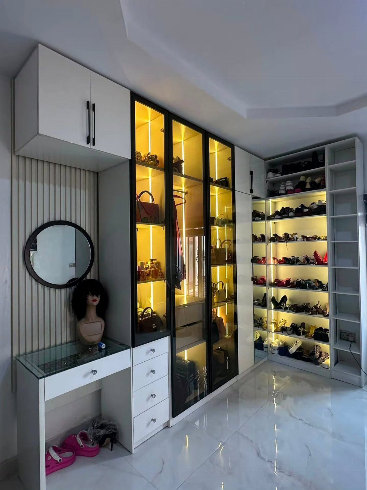 Modern walk-in closet with white dressers, illuminated glass wardrobes, and extensive shoe racks.