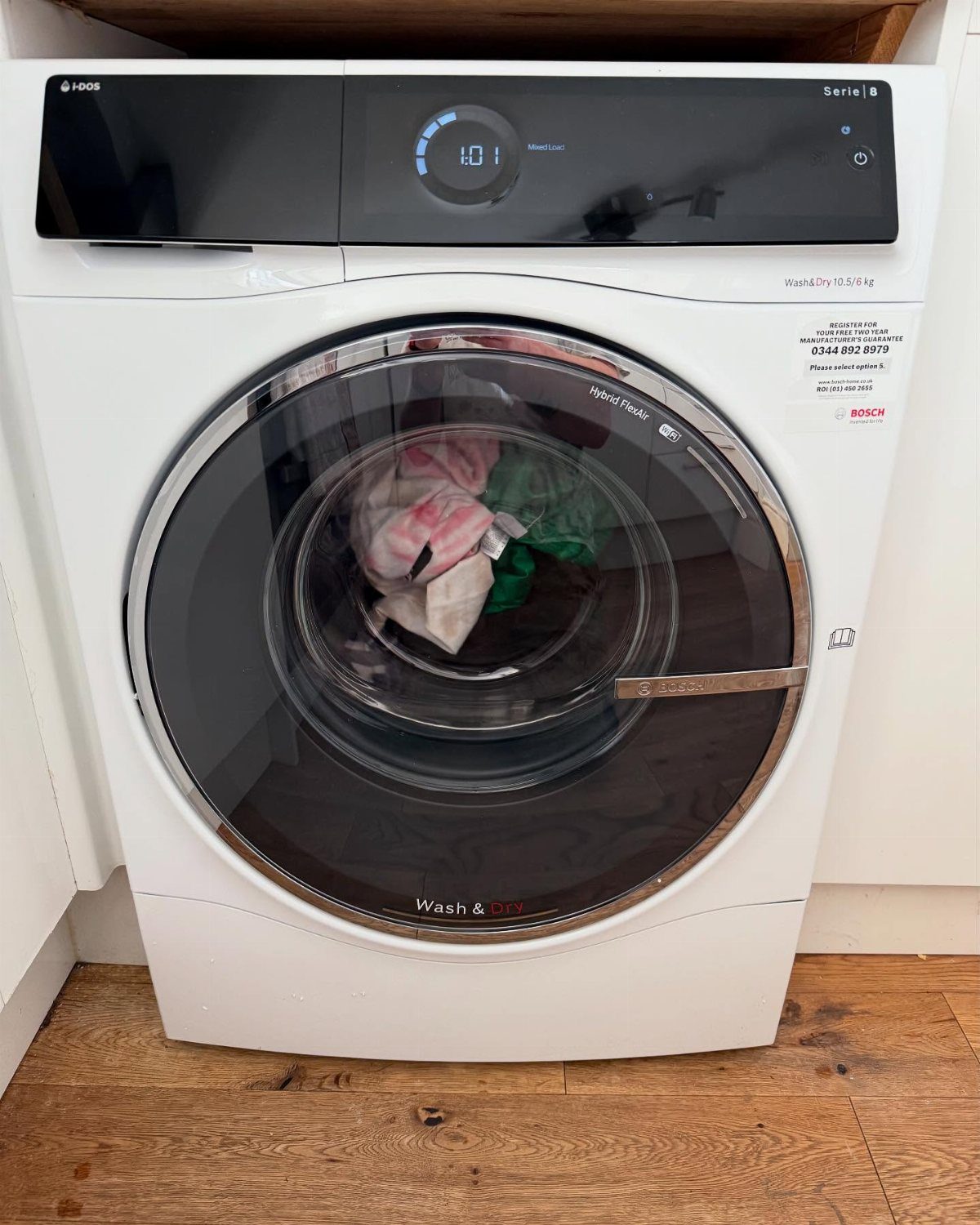 White Bosch washer-dryer combo machine with colorful clothes inside on wooden flooring.