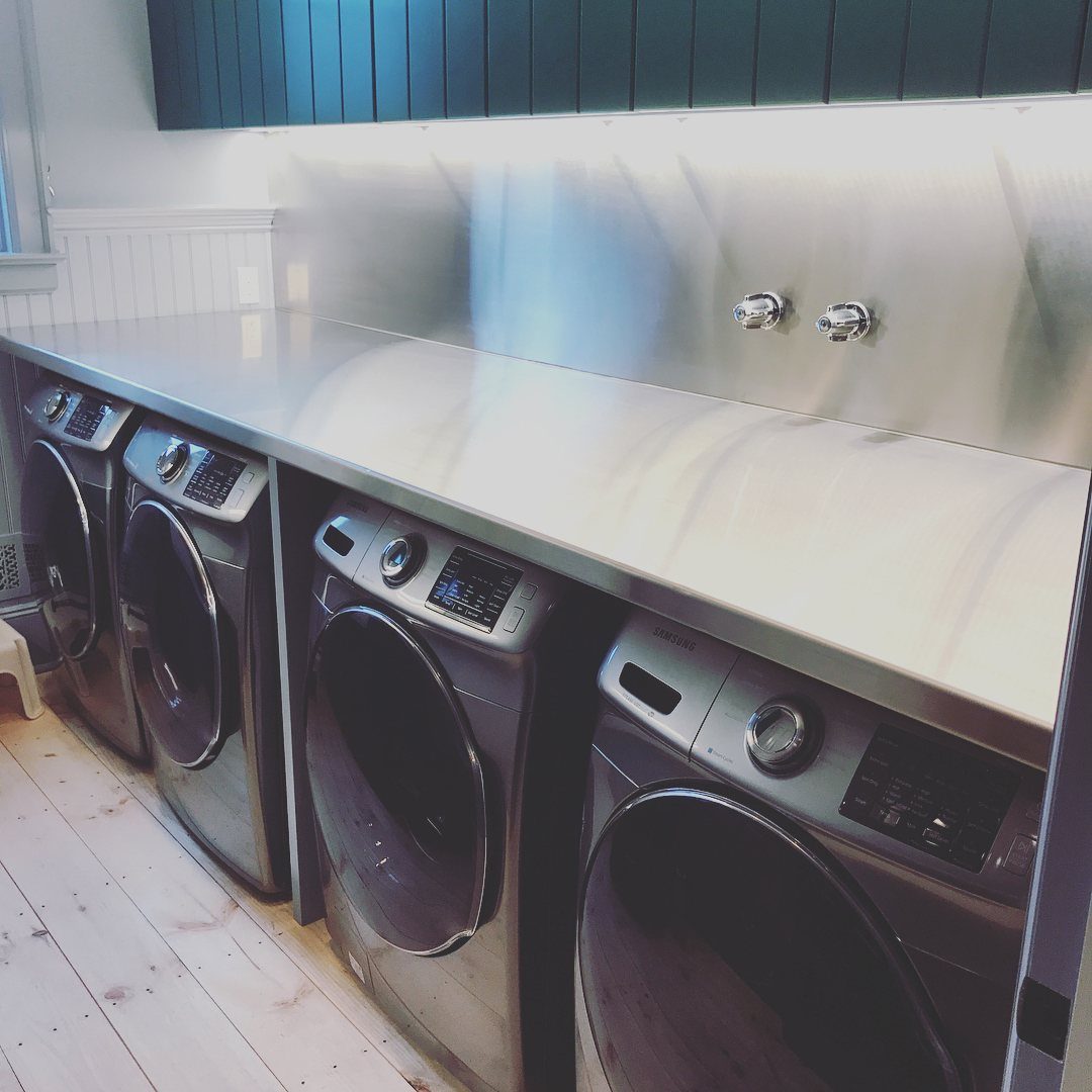 Stainless steel backsplash behind a white countertop and four front-loading washing machines.
