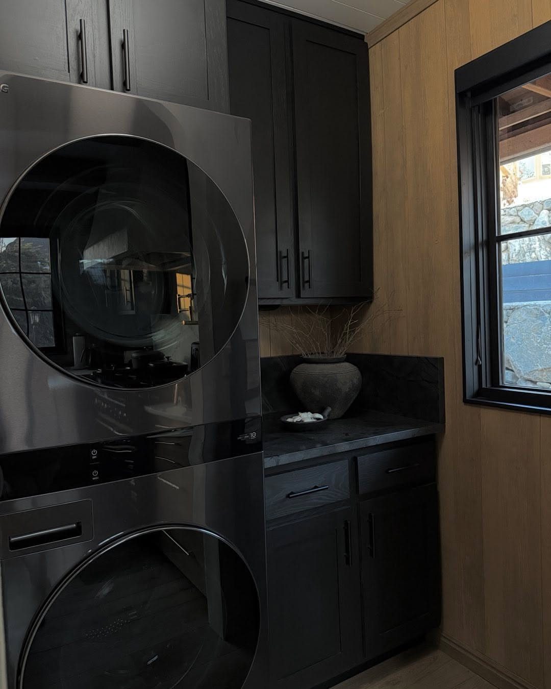 Black cabinets and stacked stainless steel laundry machines in a wood paneled room.