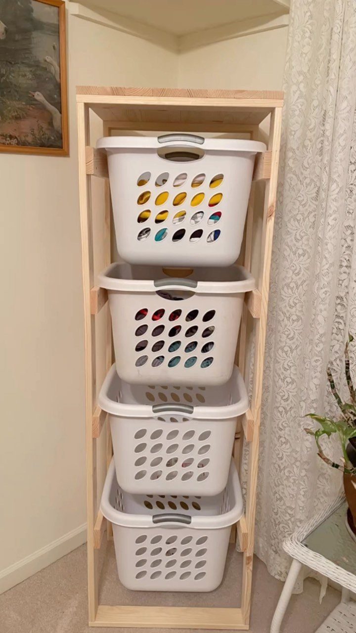 Four white laundry baskets stacked inside a light wood frame against a wall.