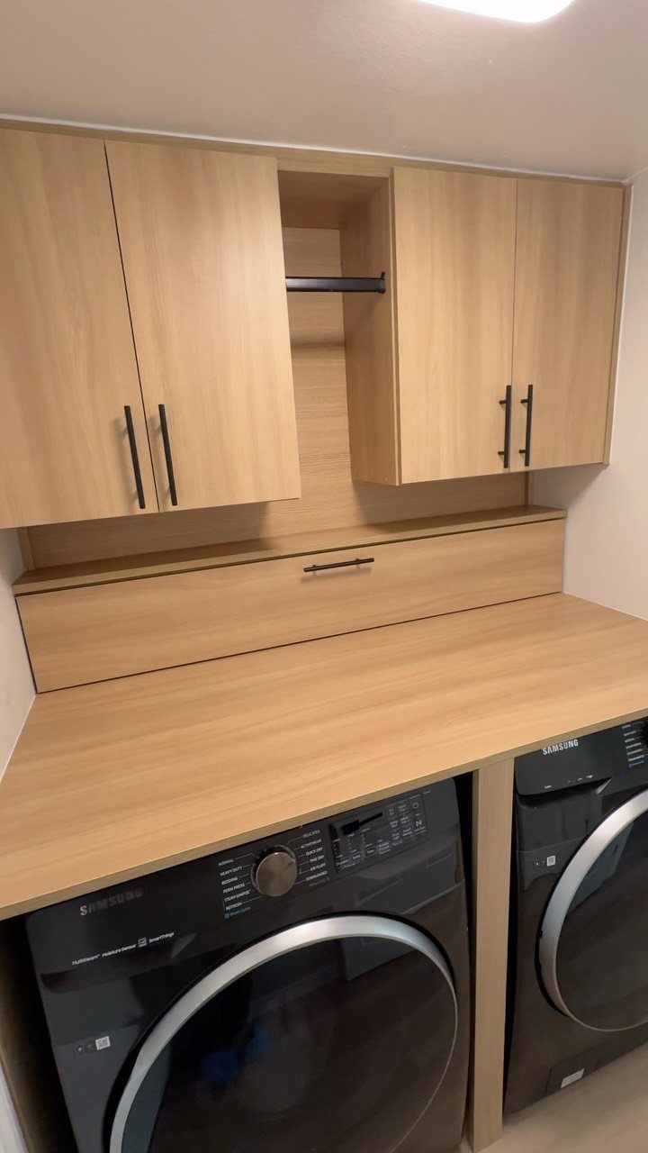 Light wood cabinets and countertop installed above two dark grey front-loading washing machines.