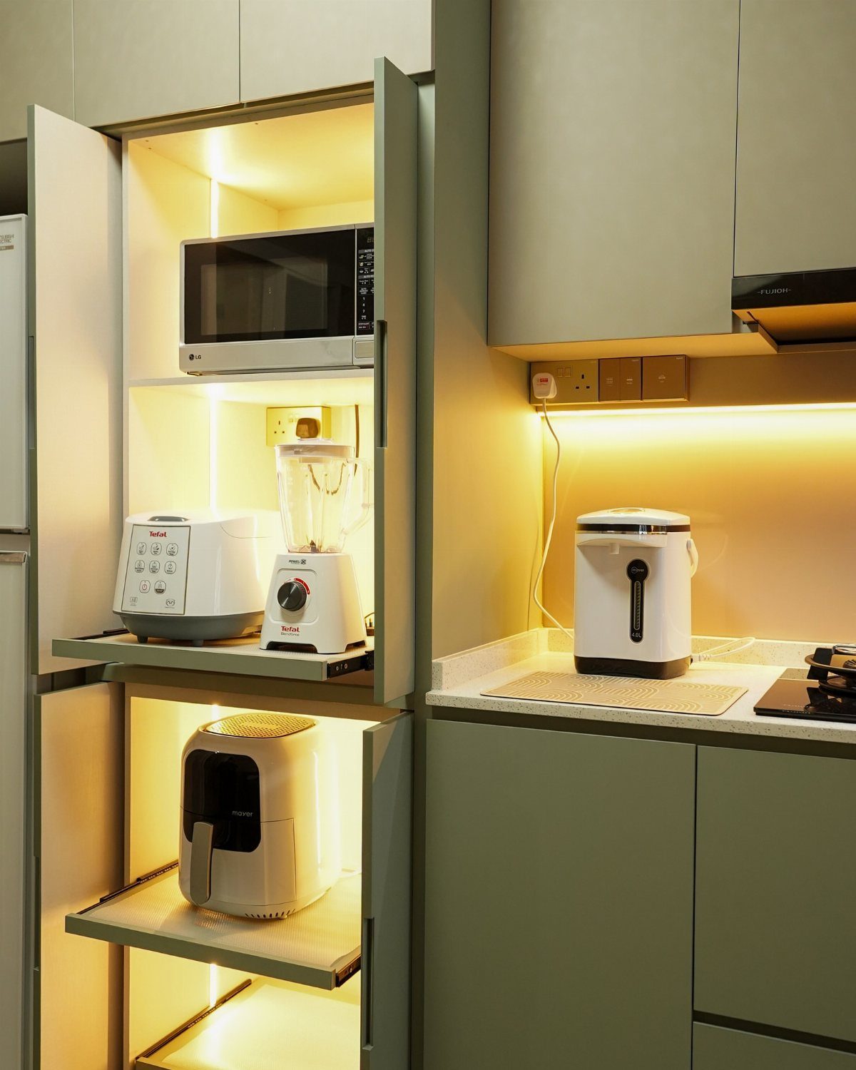Kitchen appliances stored inside a sage green cabinet with integrated warm shelf lighting.