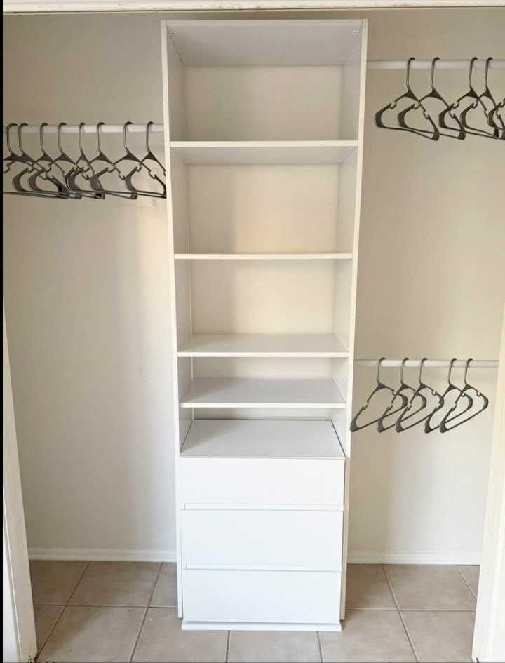 White closet organizer with central shelves, drawers, and double hanging rods with hangers.