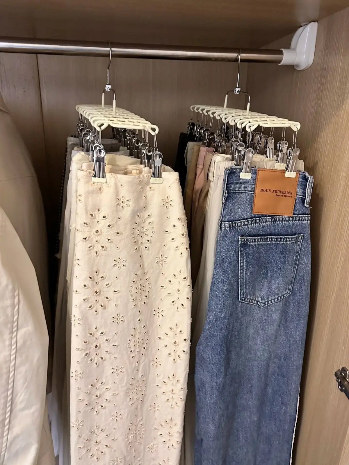 Multi-layered white hangers holding embroidered white pants and blue jeans in a closet.