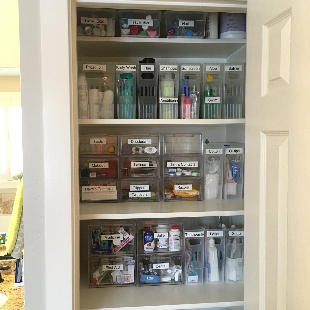 White bathroom closet shelves with labeled clear plastic bins organizing various toiletries.
