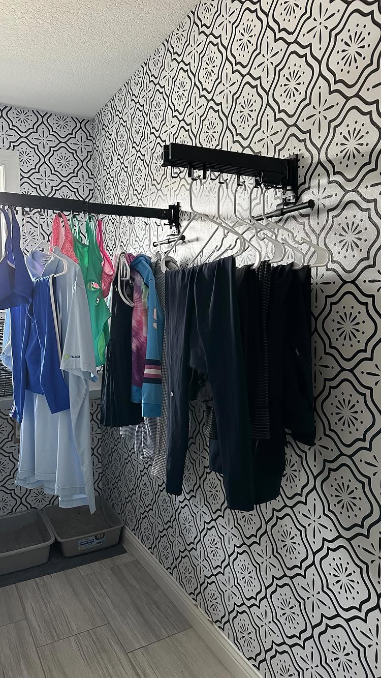 Black hanging rod with colorful clothes on hangers against patterned wallpaper.