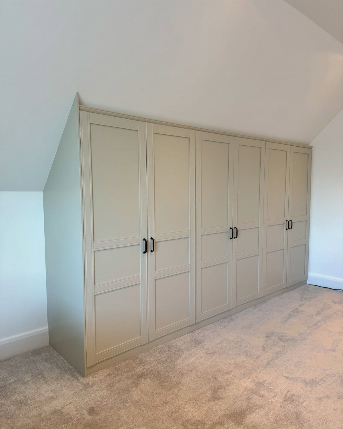 Built-in beige wardrobes installed beneath a white slanted ceiling in a carpeted room.