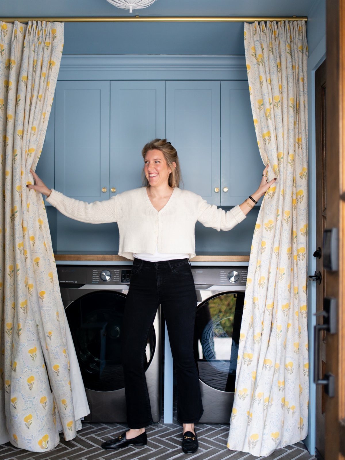 Woman opening floral curtains to reveal blue cabinets and laundry machines in a room.