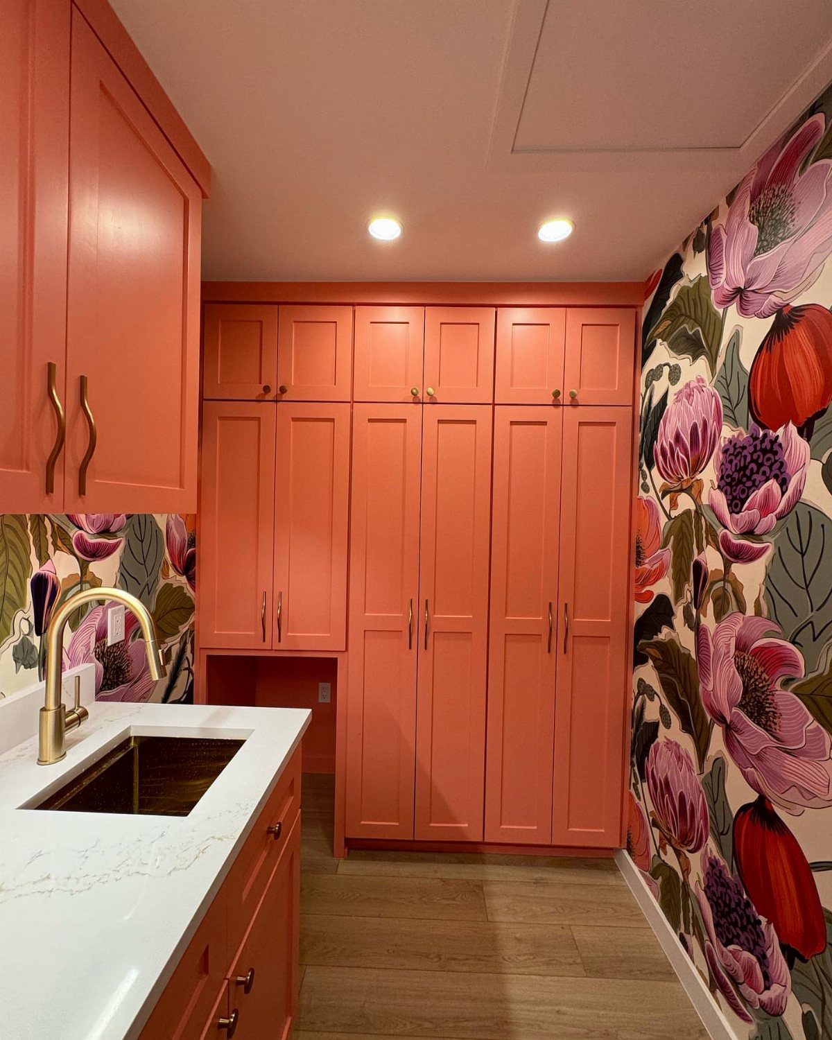 Coral cabinets and floral wallpaper in a colorful laundry room with white countertops.