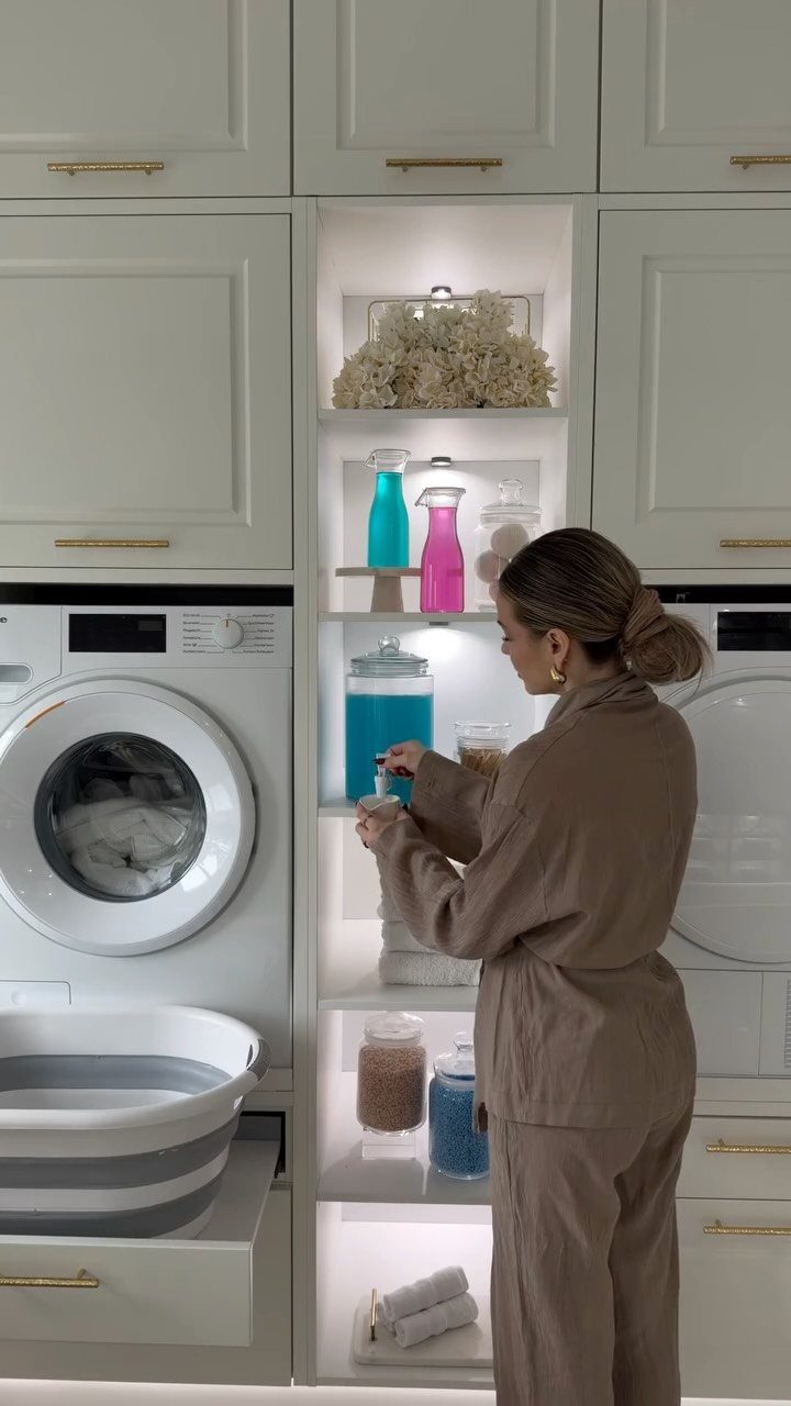 Woman reaching for detergent in illuminated white laundry room shelving next to washer