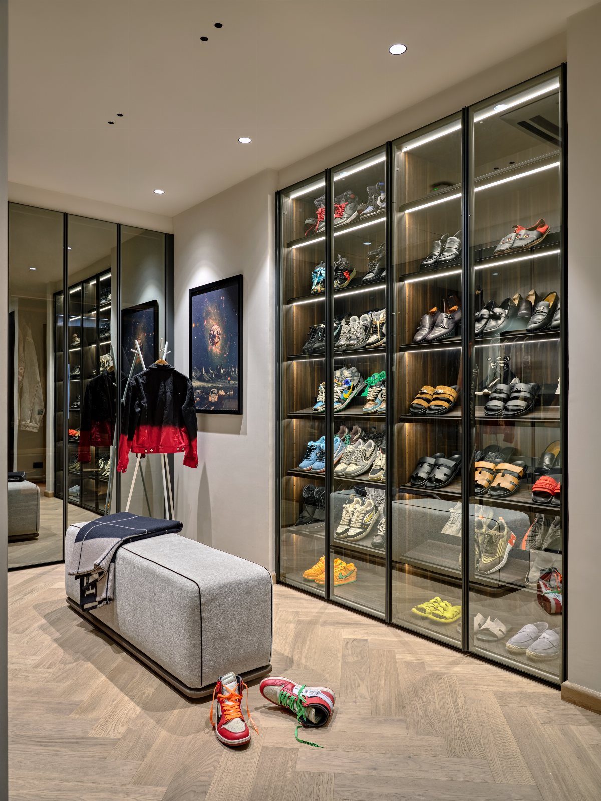 Glass-door shoe wall displaying sneakers in a modern walk-in closet with herringbone floors.