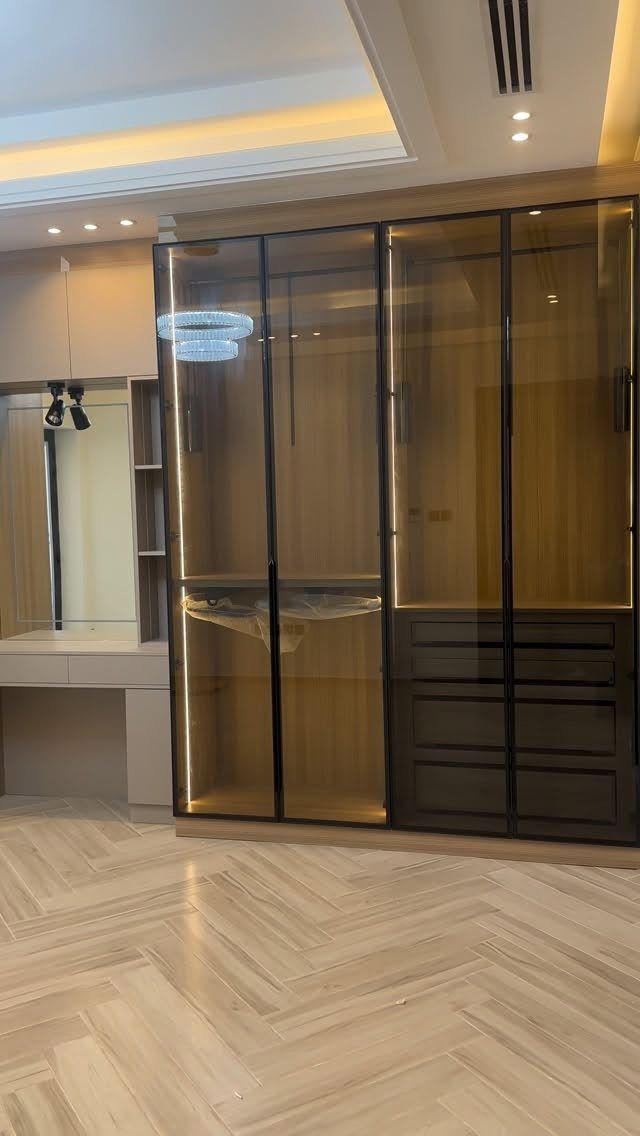 Contemporary walk-in closet with glass-door cabinets, integrated lighting, and herringbone wood floors.