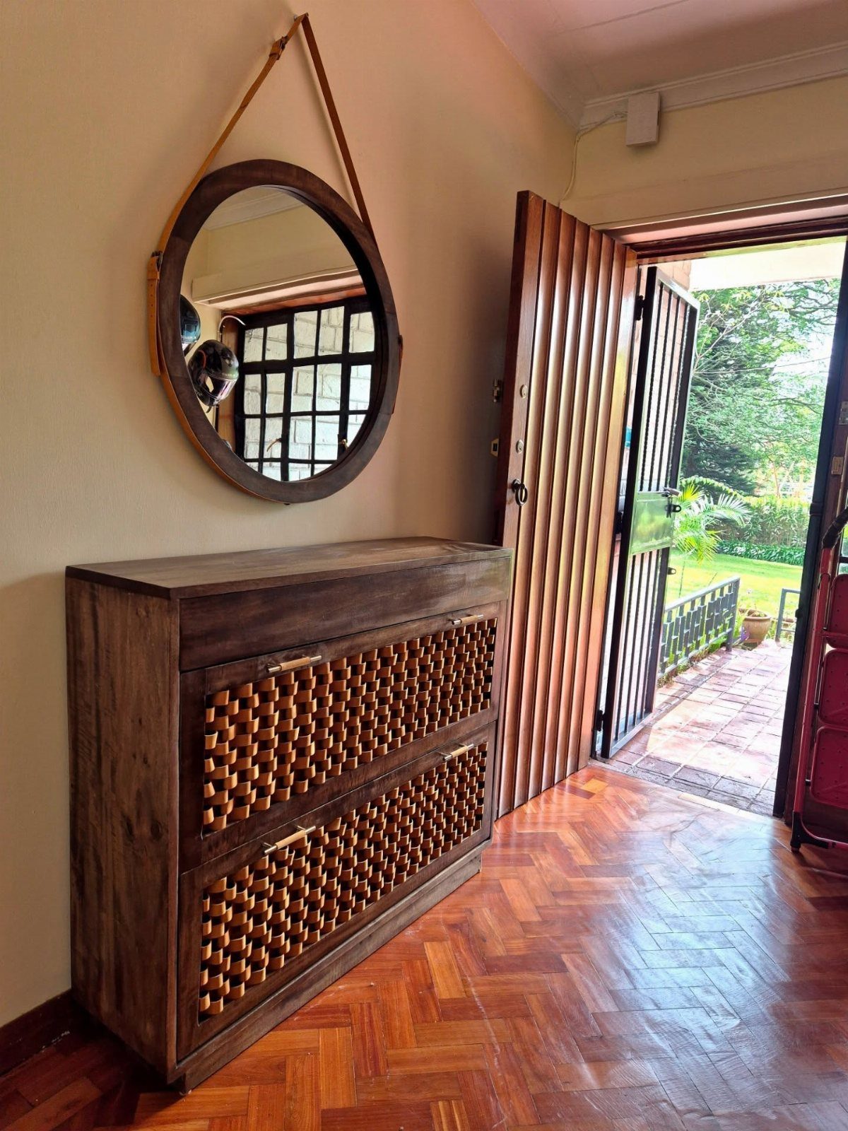 Wooden rattan cabinet and round mirror in an entryway by an open door.