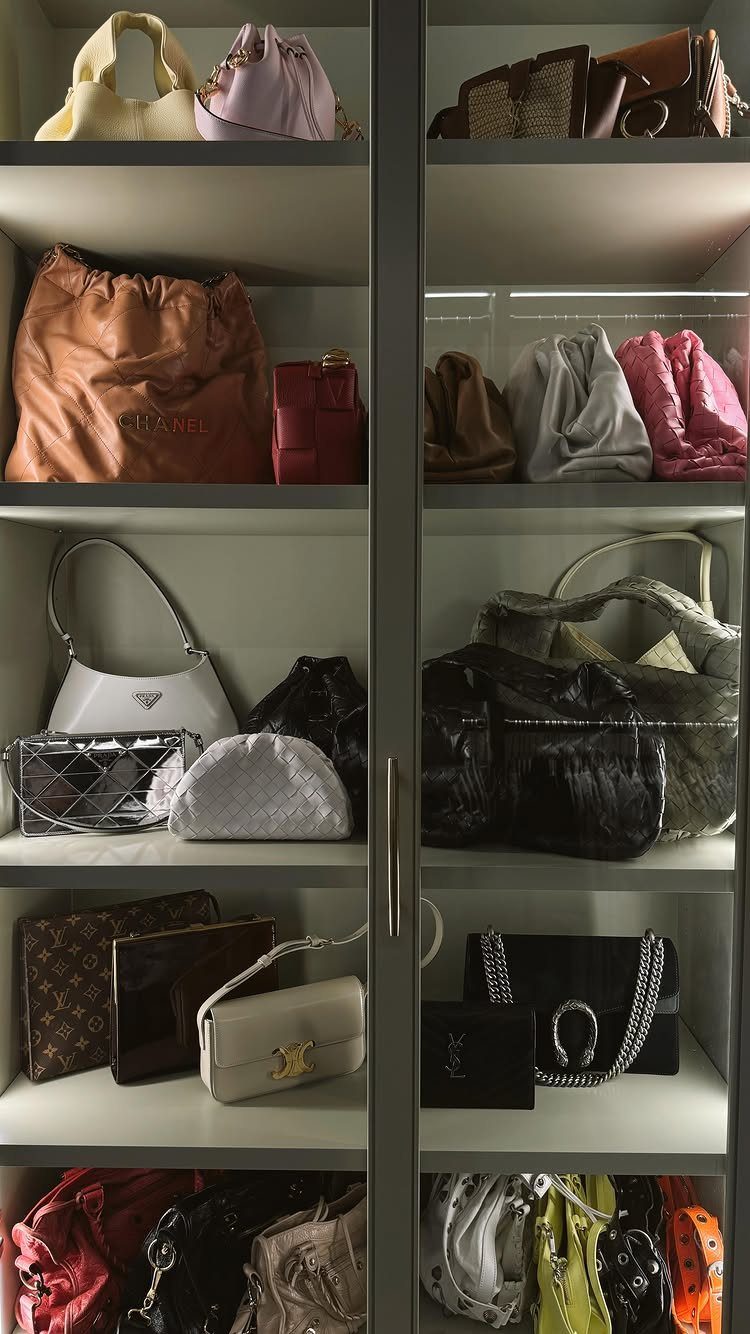 Designer handbags organized on grey shelves inside a glass-door closet.