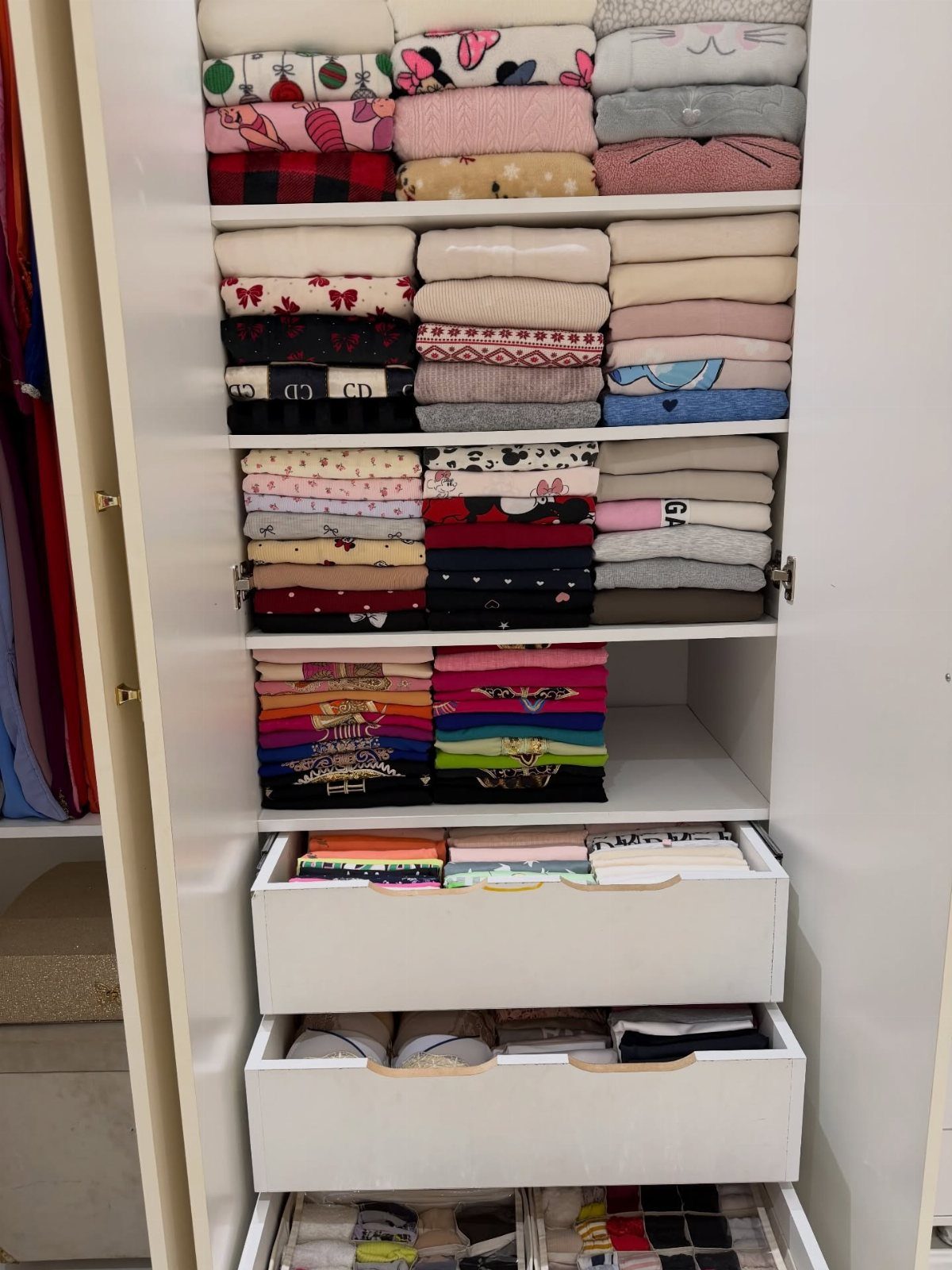 Folded clothes organized by color on white closet shelves and in drawers.