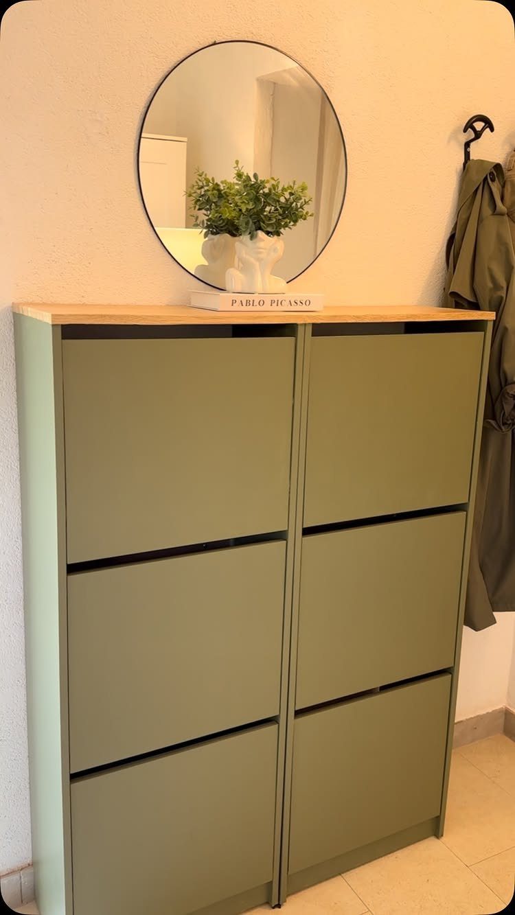 Sage green shoe storage cabinet with wooden top, round mirror and small plant.
