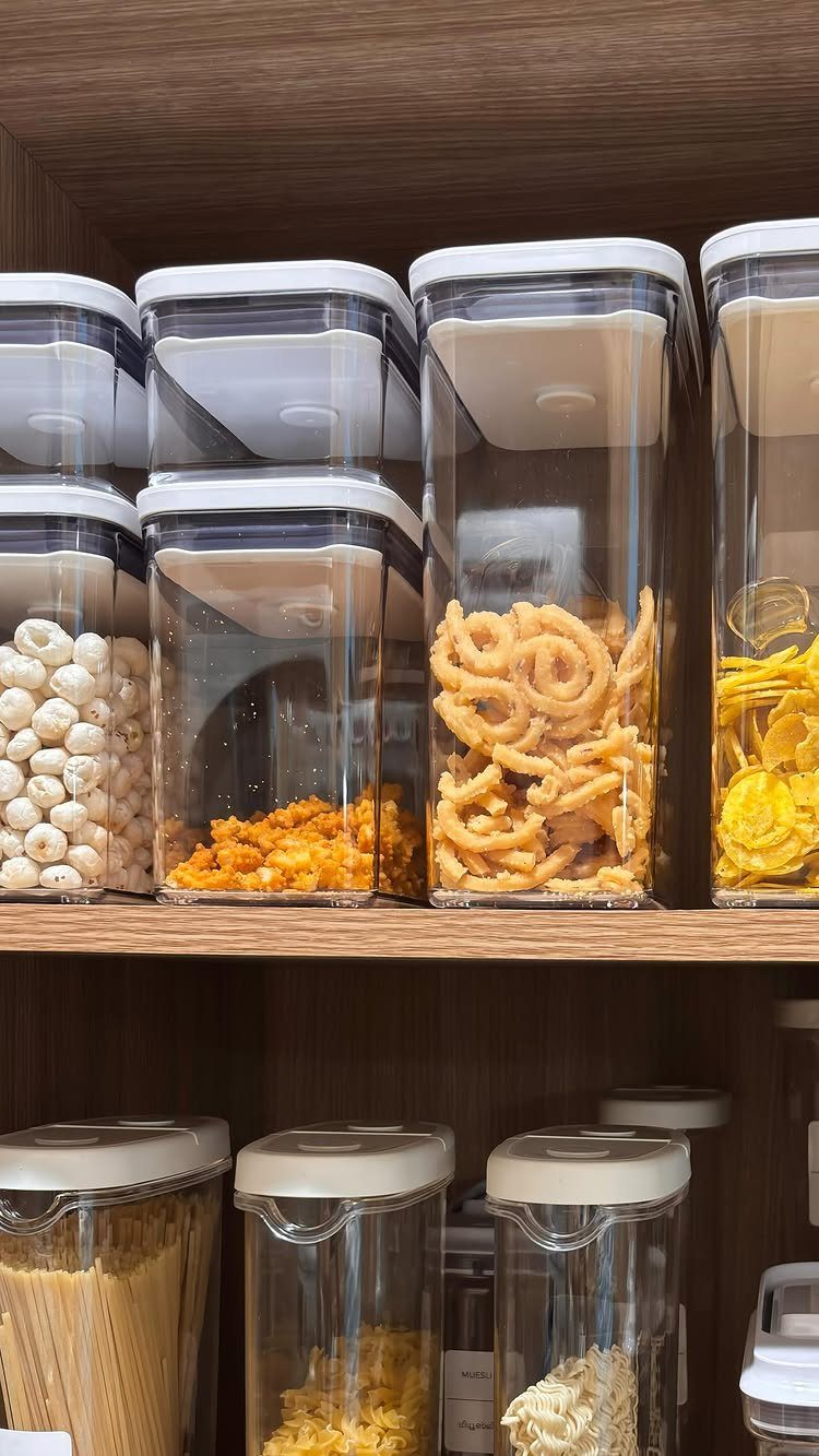 Clear airtight food storage containers filled with pasta and snacks on wooden shelves.