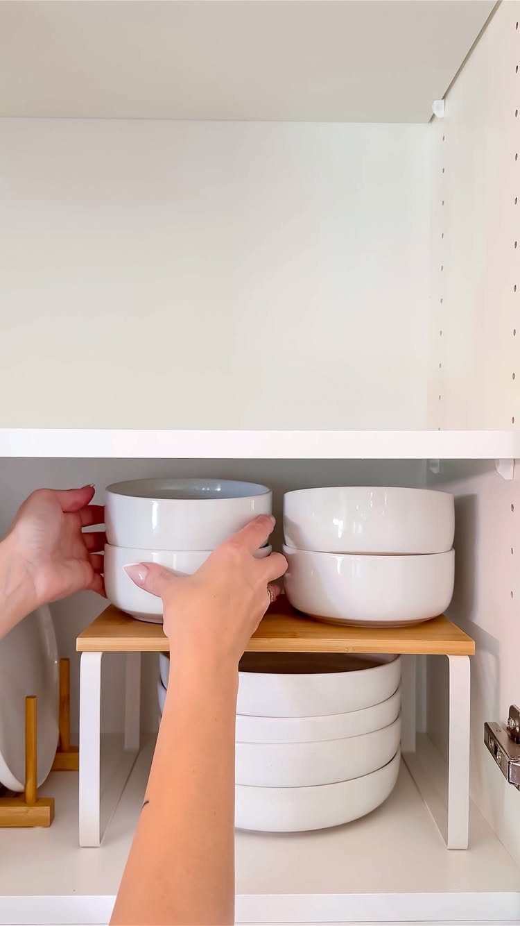 White bowls on a wooden shelf insert inside a white kitchen cabinet.