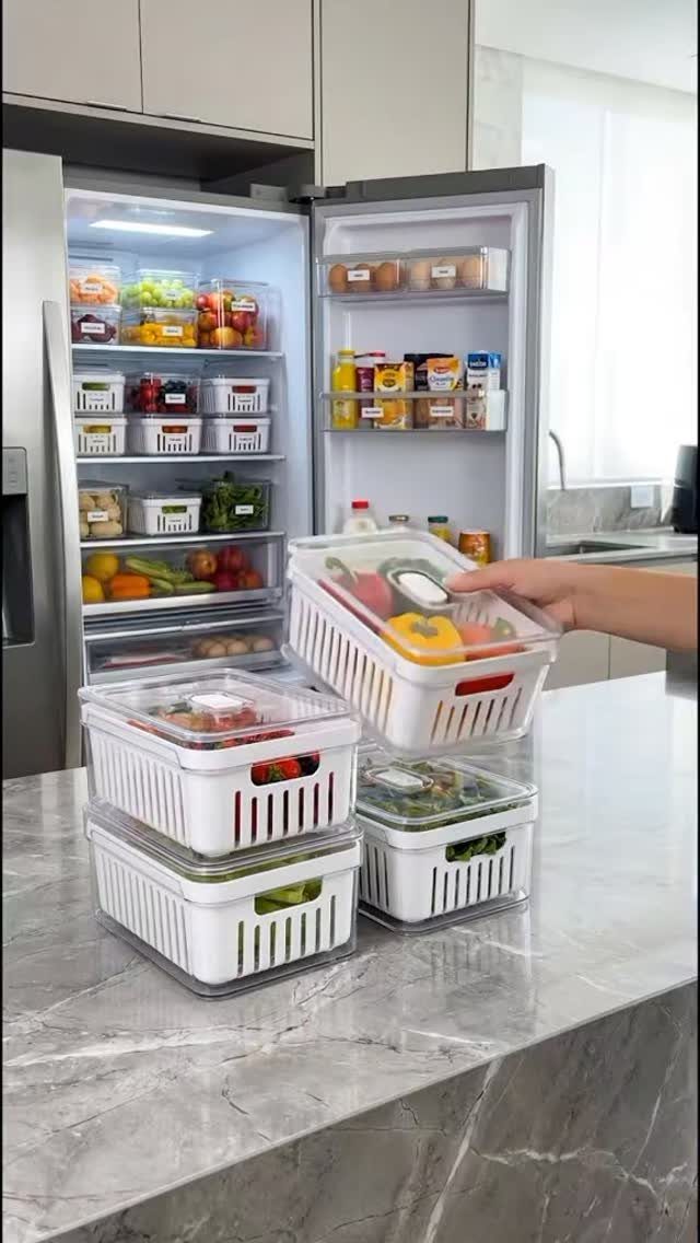 Clear stackable plastic bins with vegetables on a marble counter and open refrigerator.