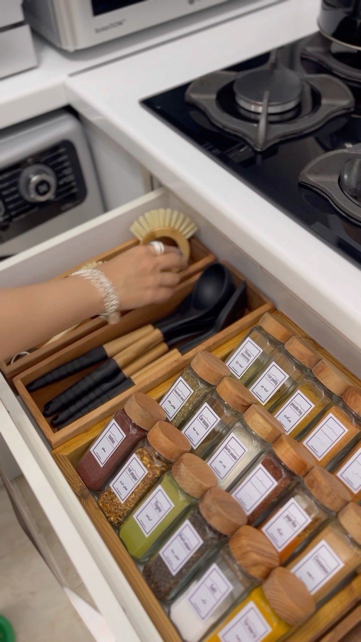 Wooden kitchen drawer organizer holding black utensils and glass spice jars with wooden lids.