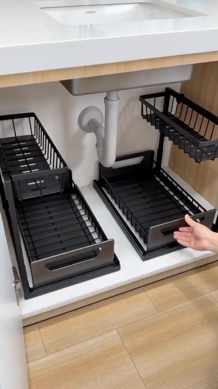 Black metal sliding organizers installed under a white kitchen sink with pull-out drawers.