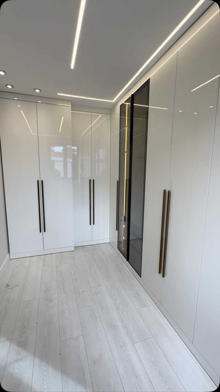 L-shaped white glossy wardrobes in a bright walk-in closet with modern ceiling lighting.