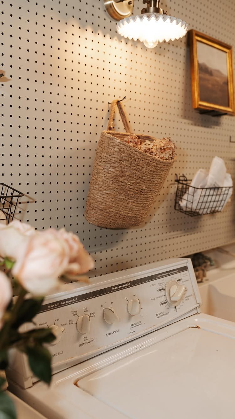 White washing machine below a white pegboard with hanging baskets and wall lighting.