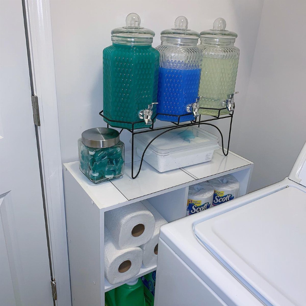 Glass beverage dispensers with colorful liquids on a white shelf next to laundry