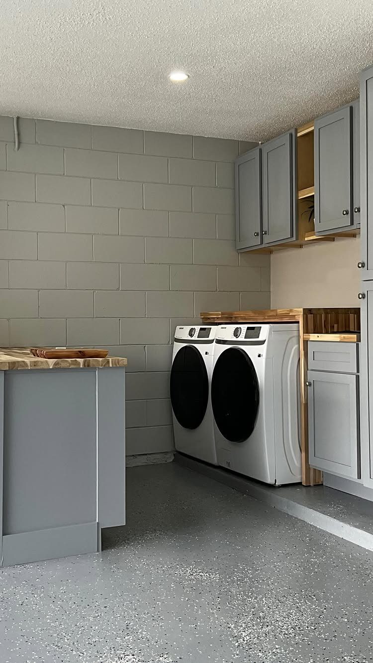 Laundry room in a garage with white machines, grey cabinets, and epoxy flooring.