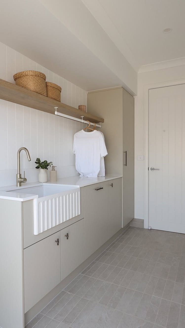 White farmhouse sink in a bright laundry room with grey cabinets and wooden shelving.