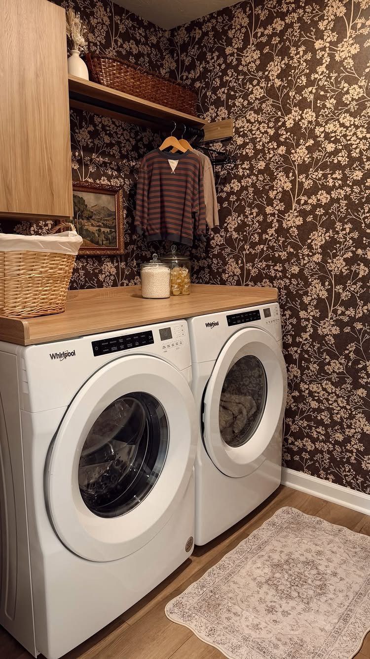 White washing machines with wooden countertop against dark brown floral wallpaper walls.