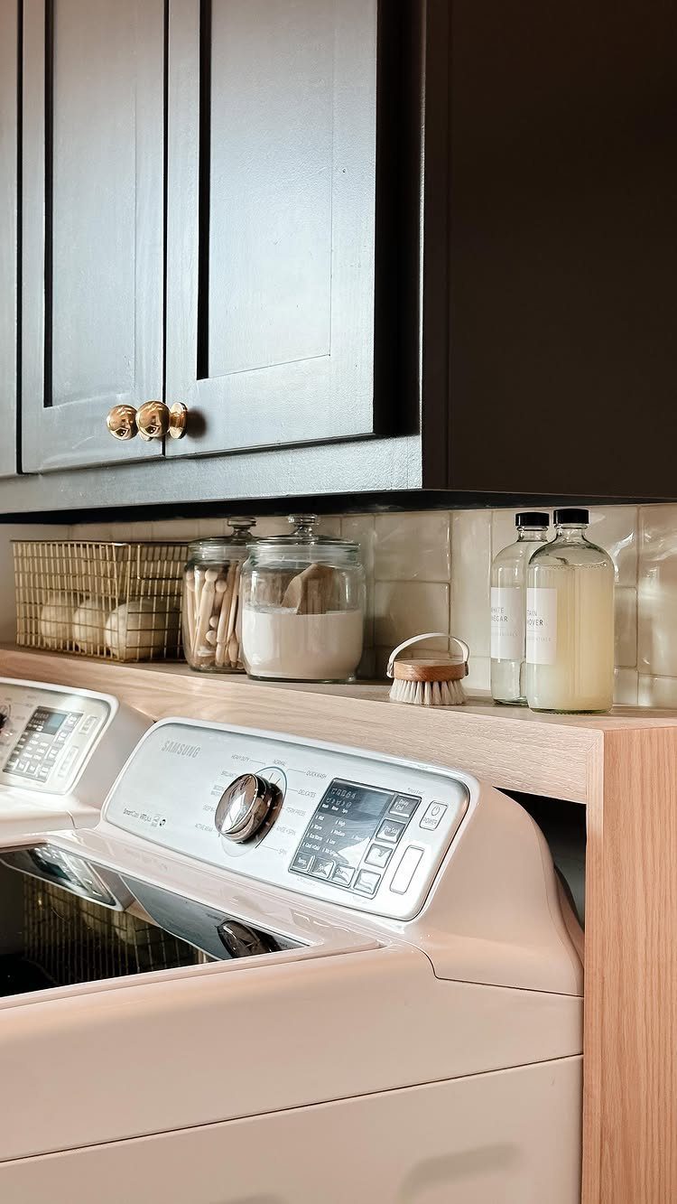 White top-loading washing machine under a wooden counter with grey cabinets above.