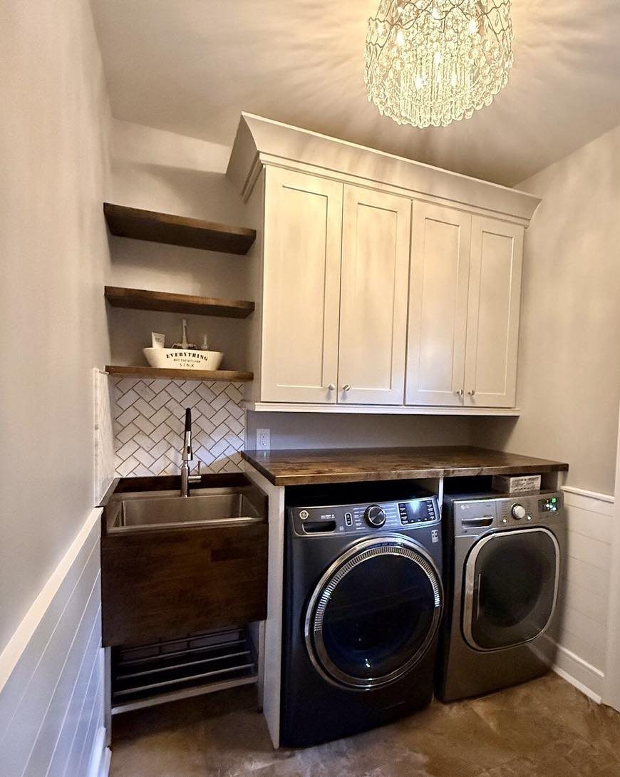 Crystal chandelier lighting a laundry room with white cabinets and dark appliances.