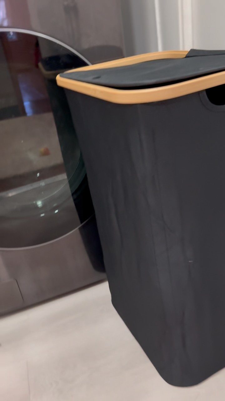 Grey fabric laundry hamper with wooden rim next to a silver washing machine.
