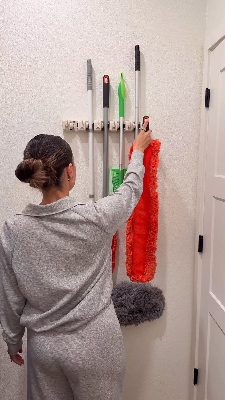 Woman reaching for a red duster on a wall-mounted cleaning tool rack.