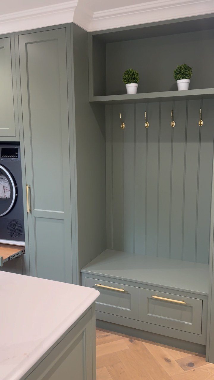 Sage green laundry room cabinetry and paneled wall with gold hooks and bench.