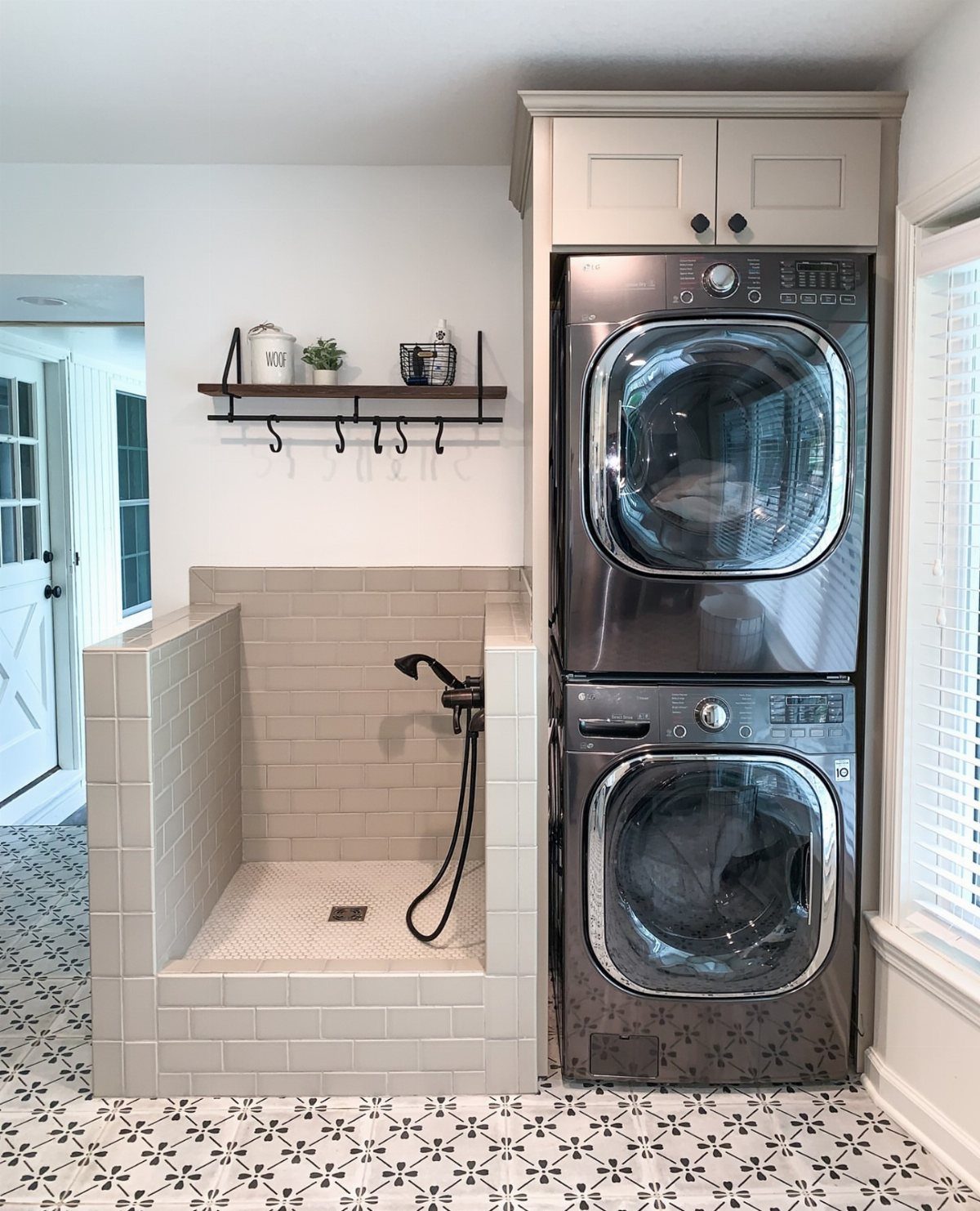 Tiled pet washing station with handheld sprayer next to stacked grey laundry machines.