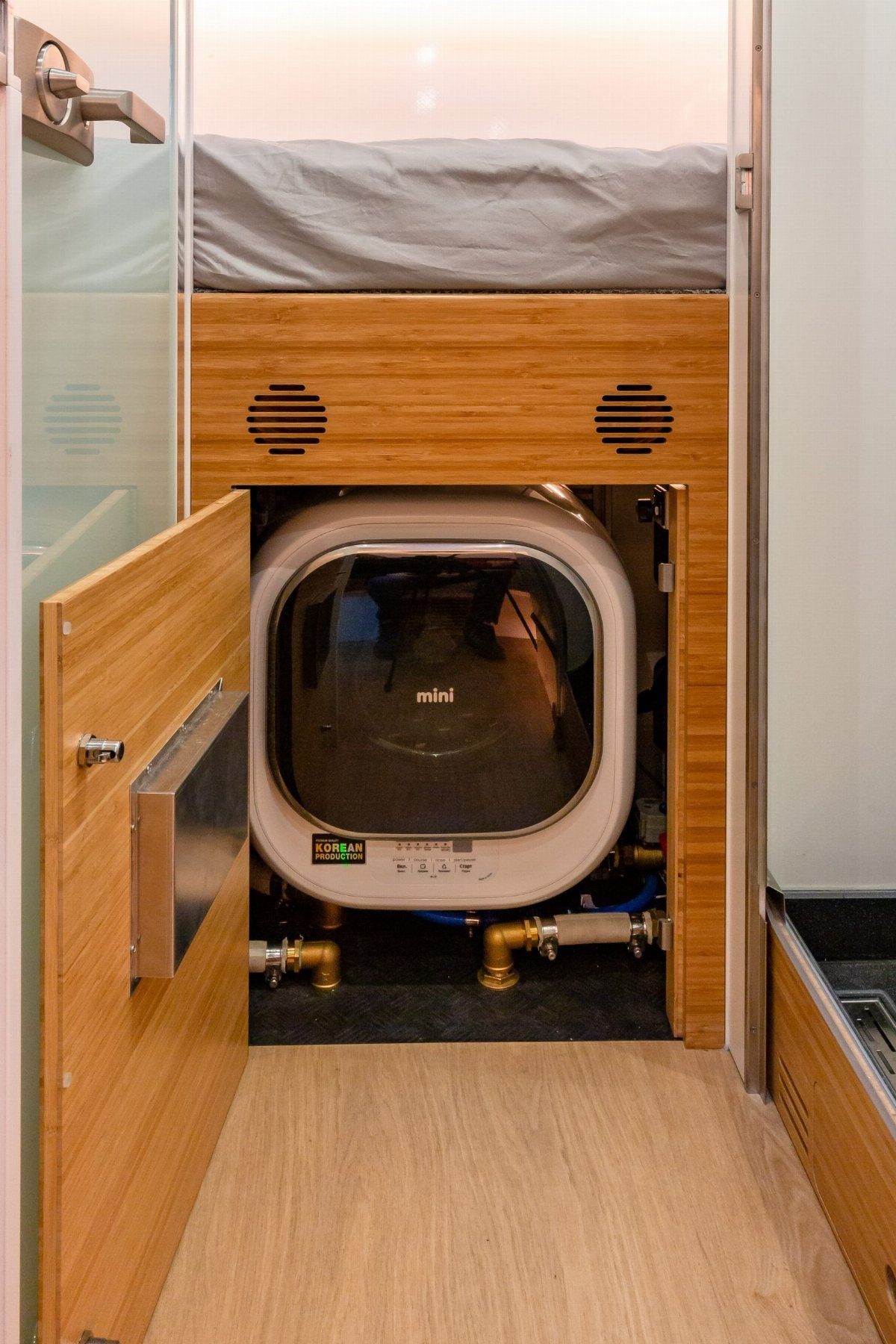 Mini washing machine installed in a wooden cabinet beneath a raised bed platform.