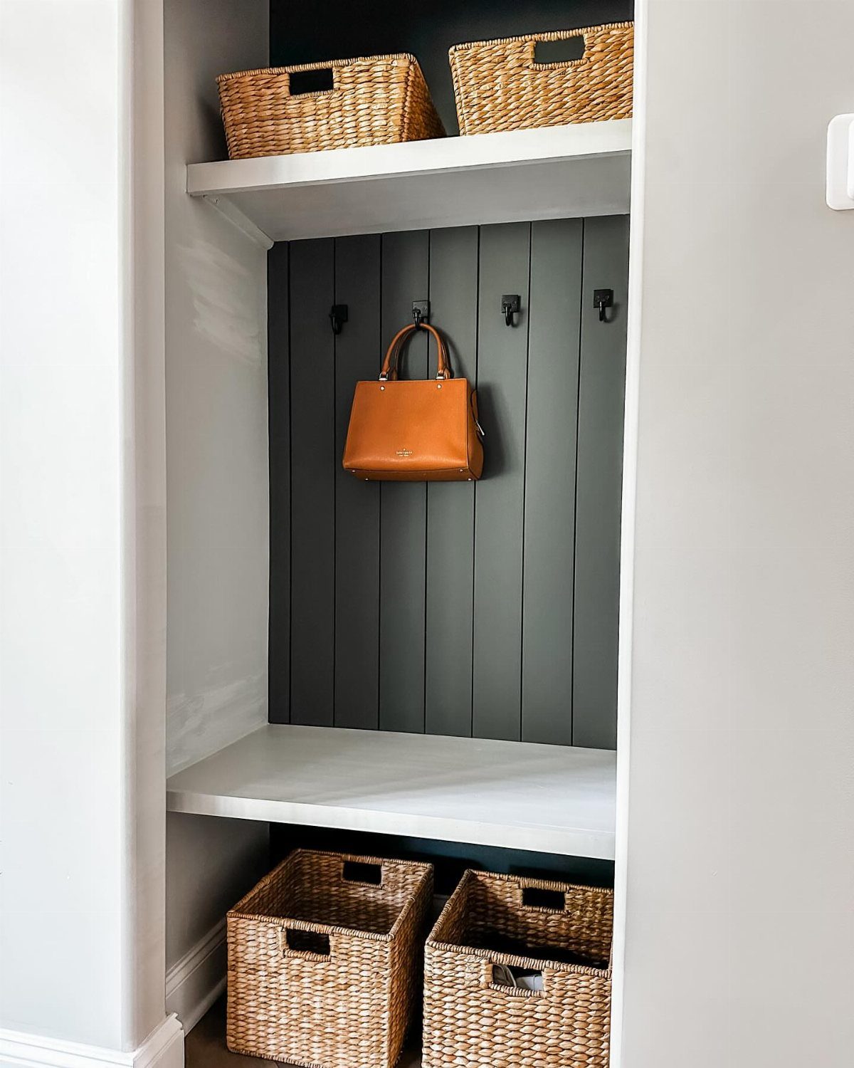 Small mudroom nook with green paneled walls, white shelving, and wicker storage baskets.