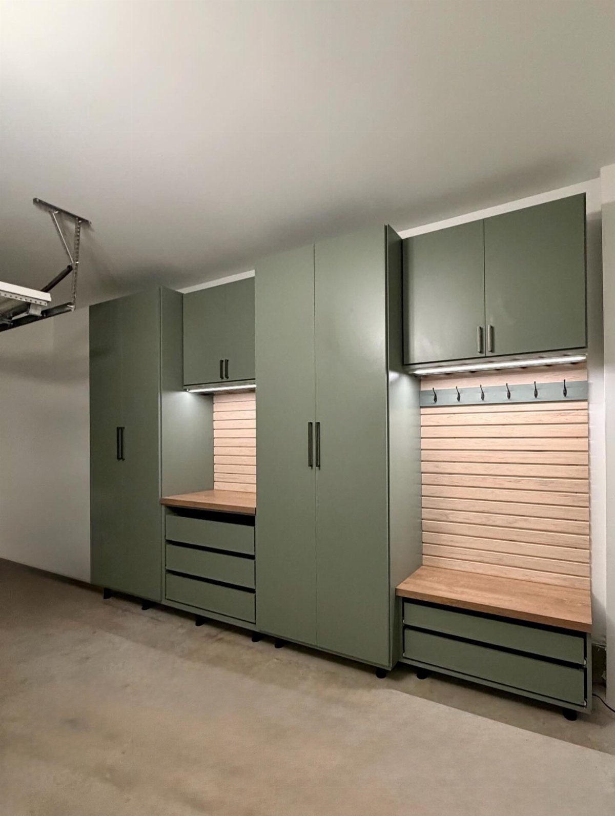 Sage green storage cabinets and lockers with wooden benches in a garage mudroom.