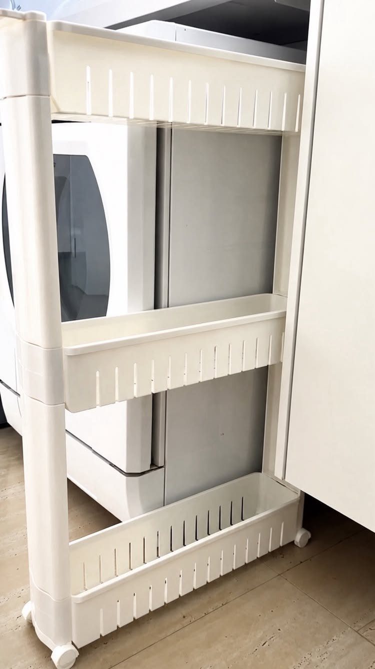 White slim rolling cart with three tiers tucked between a washer and cabinet.