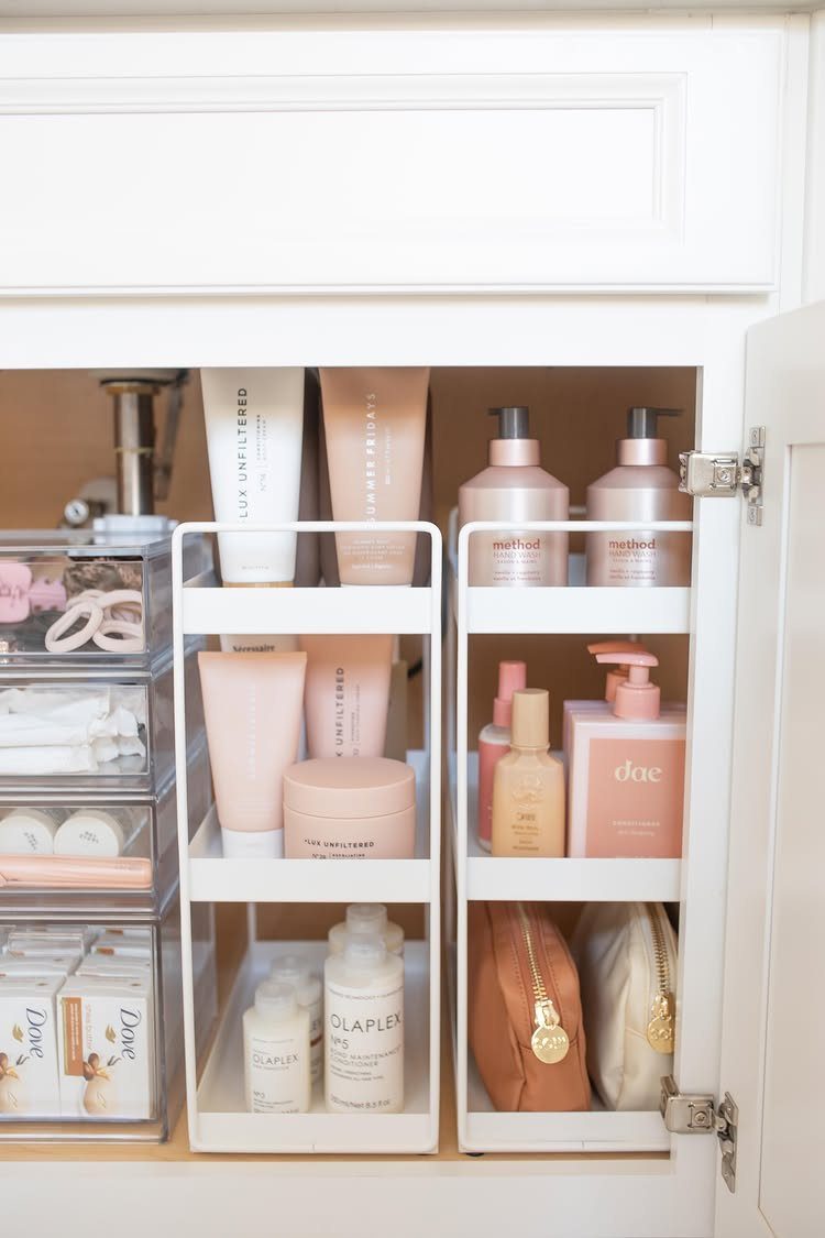 White tiered organizers and clear bins holding skincare products inside a white cabinet.