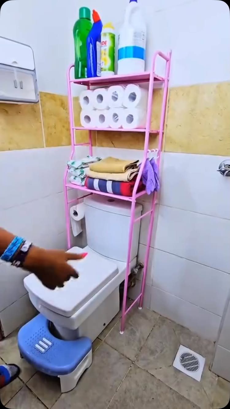 Pink over-the-toilet storage rack with cleaning supplies and towels in a small bathroom.