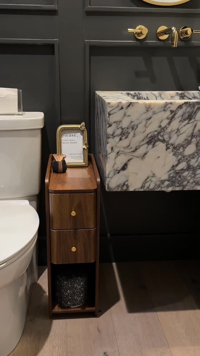 Narrow wooden storage cabinet placed between a white toilet and a marble sink.
