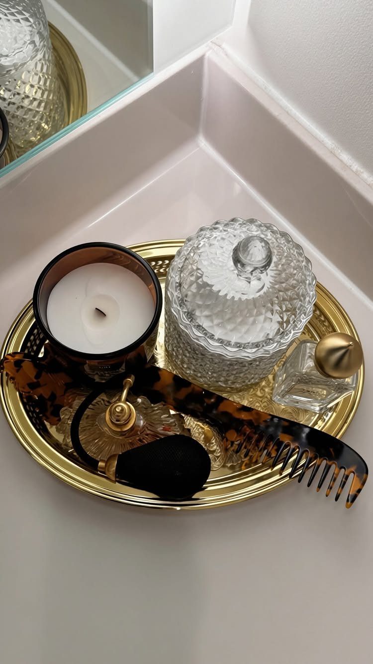 Gold oval vanity tray holding a candle, glass jar, perfume, and hair accessories.