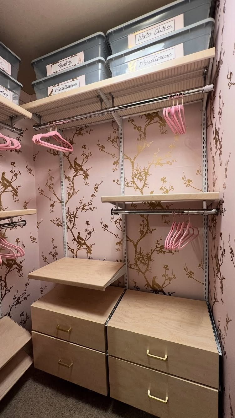 Walk-in closet with pink floral wallpaper, light wood shelves, and clear storage bins.