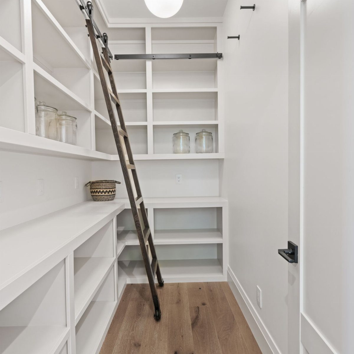 Dark wood rolling ladder on a rail in a white walk-in pantry.