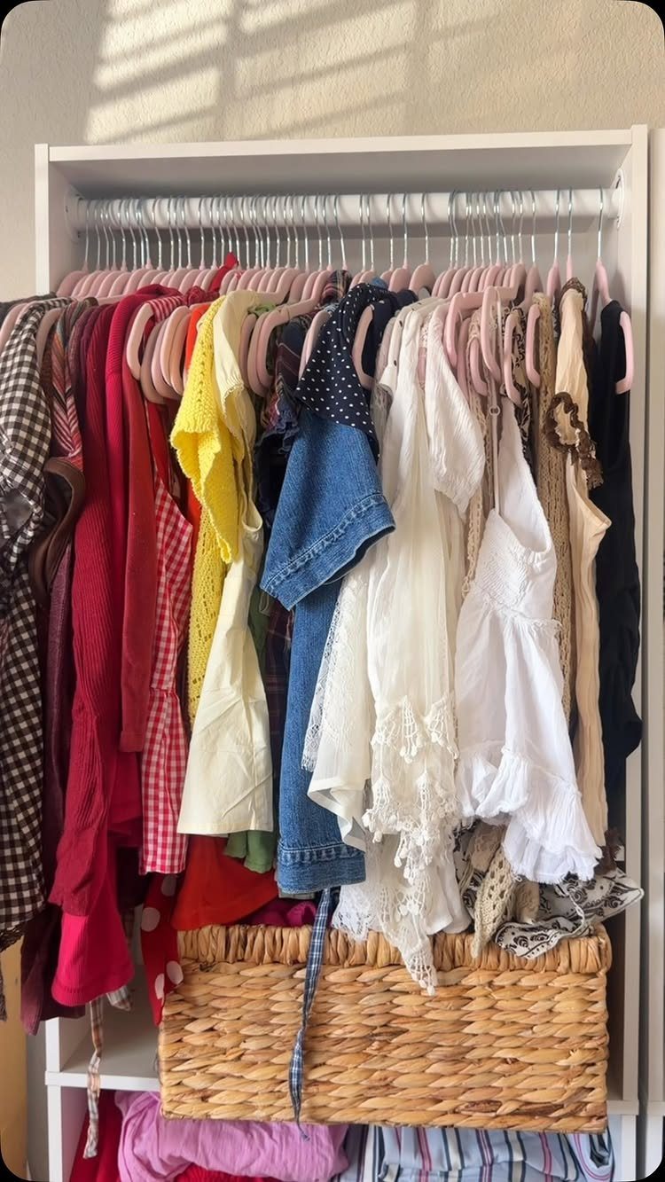 Colorful clothes on pink velvet hangers in a white reach-in closet with wicker basket.